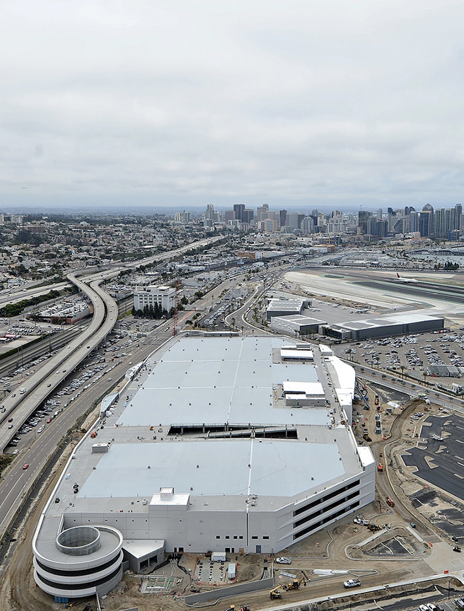 Aerial view of a large, white industrial building under construction, with a bustling cityscape and busy highways in the background, under an overcast sky.