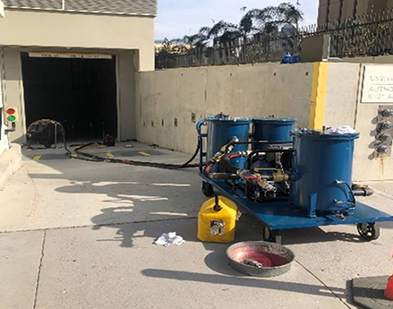 Fuel cleaning setup with blue filtration tanks and hoses on a portable cart, placed near an underground tank with a yellow gas canister and absorbent papers on the ground.