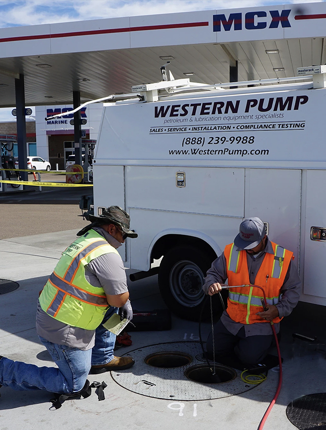Two workers in high-visibility vests performing maintenance at a gas station, with one handling a hose and the other opening an underground tank, beside a service vehicle with the logo "WESTERN PUMP".