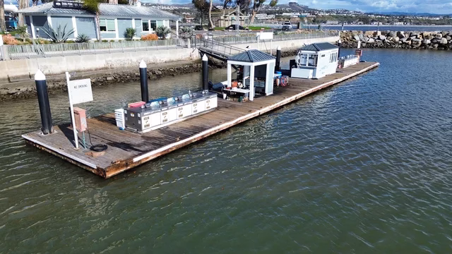 A floating dock with fuel pumps and small buildings is anchored near the shoreline at Dana Point Fuel Dock in Dana Point Marina, CA.