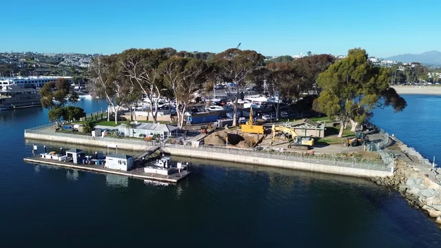 Aerial view of Dana Point Marina, CA, showing boats, trees, construction equipment, and the Dana Point Fuel Dock next to a docked barge on calm water.