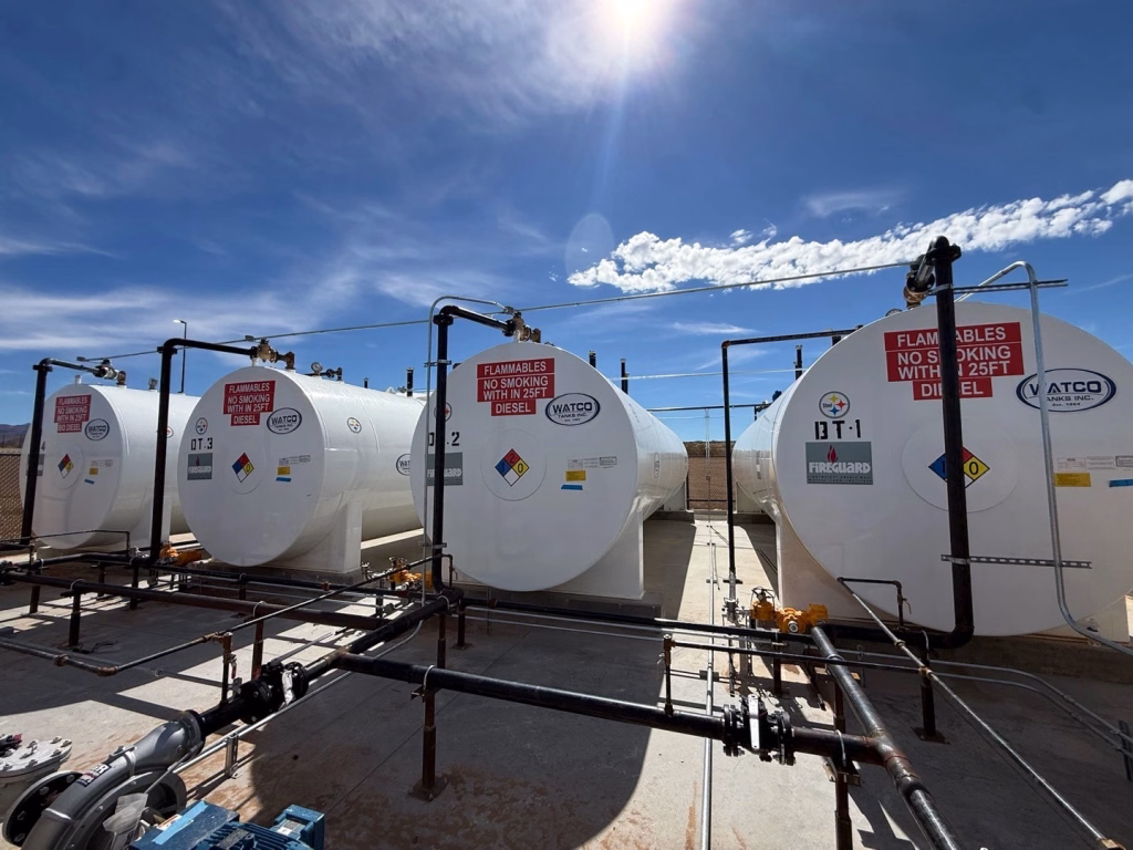 Four large white fuel storage tanks labeled for diesel stand outdoors at Love’s Travel Stop in Kingman AZ, connected by pipes with safety warnings and hazard symbols visible. The sky is mostly clear.