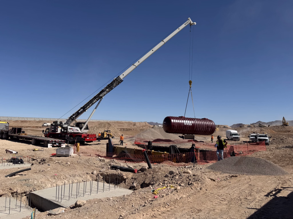 A crane lifts a large cylindrical tank over a construction site near Love’s Travel Stop in Kingman AZ, with workers and vehicles nearby under a clear blue sky.